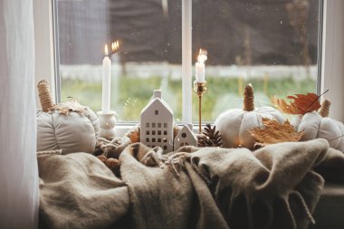Cozy Autumn. Stylish pumpkin pillows, fall leaves, candle, lights and cute buildings decoration on brown scarf on windowsill. Autumn hygge, fall home decor. Happy Thanksgiving