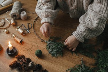 Eller rahat süveter içinde Noel çelengi yaparken köknar dalları, çam kozalakları, mum, sicim, ahşap masada çanlar, yakın çekim. Üst Manzara. Kış tatili hazırlıkları, atmosferik zaman