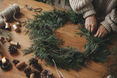 Eller rahat süveter içinde Noel çelengi yaparken köknar dalları, çam kozalakları, sicim, ahşap masada çanlar, kapat. Kış tatili hazırlıkları, atmosferik zaman