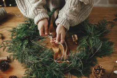 Noel kırsal çelengi hazırlıyorum. Kurdele tutan eller, sedir dalları, çam kozalakları ile ahşap masa üzerinde çelenk yapmak, atmosferik festival odasında çanlar. Kış tatili hazırlıkları