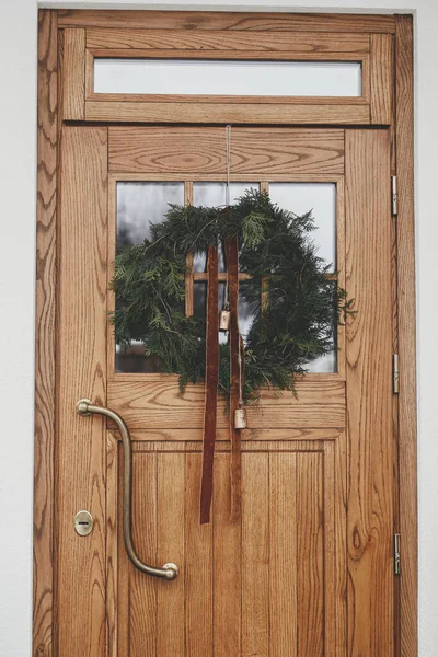 别致的圣诞花环 木门上有老式的铃铛和带子 屋外有冷杉树和灯笼 现代农舍立面冬季假日装饰 — 图库照片