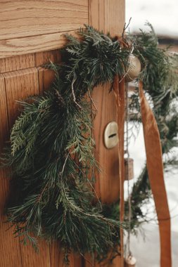 Pirinç sapında eski çan ve kurdele asılı olan şık bir Noel rustik çelengi ahşap ön kapıya yakın. Kış kırsal tatilleri için modern eko çiftlik evi dış dekorasyonu