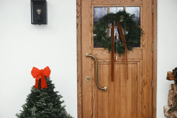 别致的圣诞花环 带有老式的铃铛和缎带 挂在木制前门 冷杉树上 房门口有红色的蝴蝶结 寒假用现代最小农舍外饰 — 图库照片