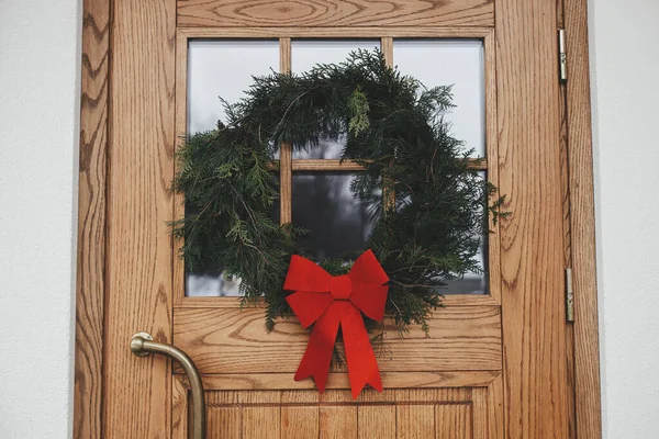 在房屋入口处的木门上有一个带红色蝴蝶结的时髦的圣诞乡村花环 冬季度假装饰的现代农舍外观 圣诞快乐 节日快乐 — 图库照片