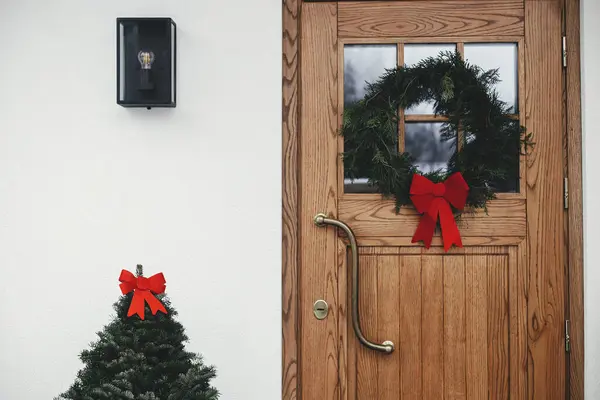 风格别致的圣诞乡村花环 红色的蝴蝶结挂在木制前门 冷杉树上 红色的蝴蝶结挂在房屋入口 寒假用现代最小农舍外饰 — 图库照片