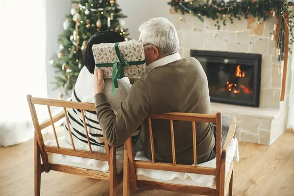Christmas present surprise. Happy senior family exchanging christmas gift and enjoying cozy fireplace. Beautiful elderly couple hugging and relaxing in stylish christmas decorated living room