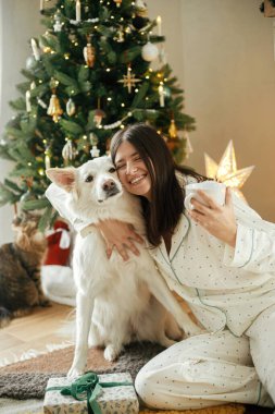 Sıcak bir Noel sabahı. Pijamalı güzel bir kadın sıcak çay tutuyor ve şık bir salonda şık bir ağaçta sevimli bir köpeğe sarılıyor. Mutlu Noeller! Hayvanlarla kış tatili