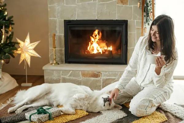 Cozy christmas morning. Beautiful woman in stylish pajamas holding warm tea and relaxing with cute  dog at fireplace in festive decorated living room. Merry Christmas! Winter holidays with pet