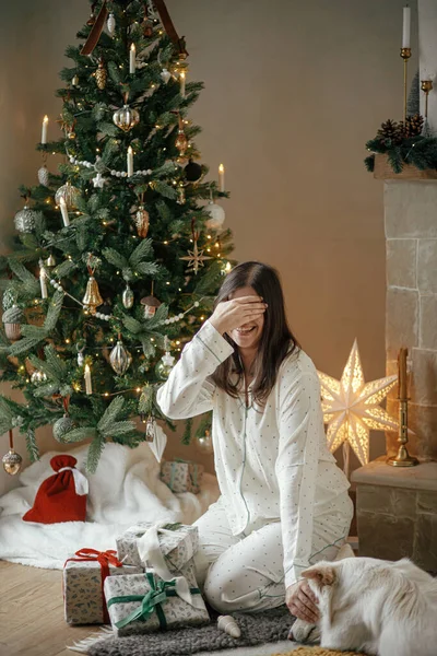 Christmas morning surprise. Beautiful woman in pajamas and cute white dog closing eyes before stylish christmas presents at decorated tree in festive living room. Merry Christmas!