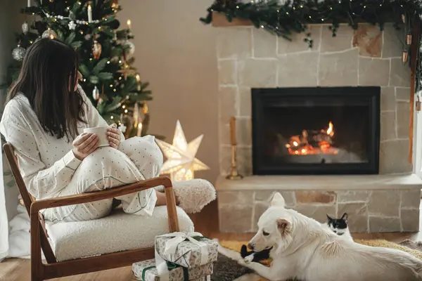 Cozy christmas morning. Beautiful woman in stylish pajamas relaxing with warm tea and cute dog at fireplace in festive decorated living room. Merry Christmas! Winter holidays with pet