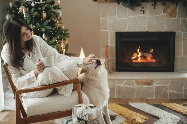 Cozy christmas morning. Beautiful woman in stylish pajamas relaxing with warm tea and cute dog at fireplace in festive decorated living room. Merry Christmas! Winter holidays with pet