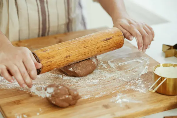 Hands kneading gingerbread dough on wooden board with rolling pin, golden metal cutters, cooking spices in modern white kitchen. Woman making christmas gingerbread cookies