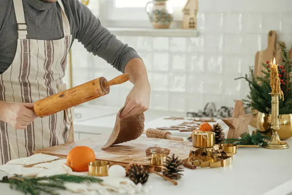 Man making christmas gingerbread cookies in modern white kitchen. Hand kneading gingerbread dough on wooden board with flour, rolling pin, golden metal cutters, cooking spices on countertop