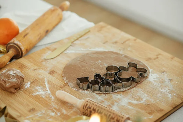 Making christmas gingerbread cookies. Gingerbread dough with festive metal cutters on wooden board with flour, cooking spices, festive decorations in modern white kitchen