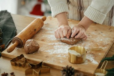 Noel kurabiyesi yapan bir kadın. Elleri zencefilli ekmek hamurunu oklavayla yoğurur, altın kesiciler, yemek baharatları, kırsal masada süslü Noel ağacına karşı şenlikli süslemeler