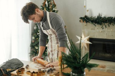 Noel kurabiyesi yapan bir adam. Elleri zencefilli ekmek hamurunu oklavayla yoğurur, altın kesiciler, yemek baharatları, kırsal masada süslü Noel ağacına karşı şenlikli süslemeler