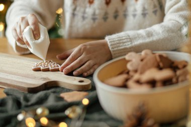 Eller, Noel kurabiyelerini kırsal ahşap masa üzerindeki kremayla süslüyor. Atmosferik Noel gelenekleri. Kurabiyeleri şekerli süslemek. Aile zamanı.