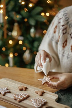 Elleri zencefilli kurabiyeleri altın ışıkların arkasındaki ahşap masa üzerindeki kremayla süslüyor. Atmosferik Noel gelenekleri. Kurabiyeleri şekerli süslemek