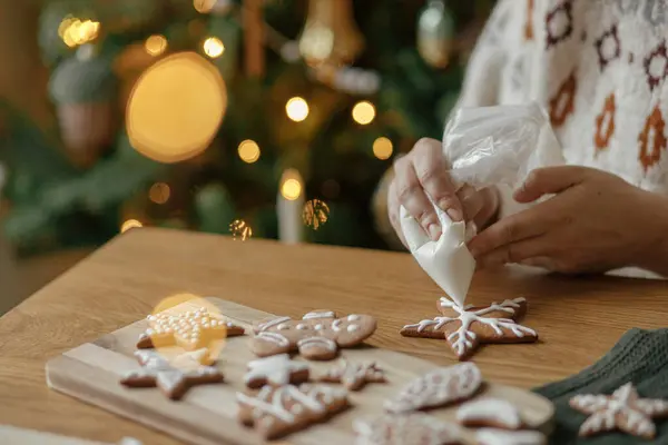 Hands decorating christmas gingerbread cookies with icing on rustic wooden table on background of golden lights. Atmospheric Christmas holiday traditions. Decorating cookies with sugar frosting