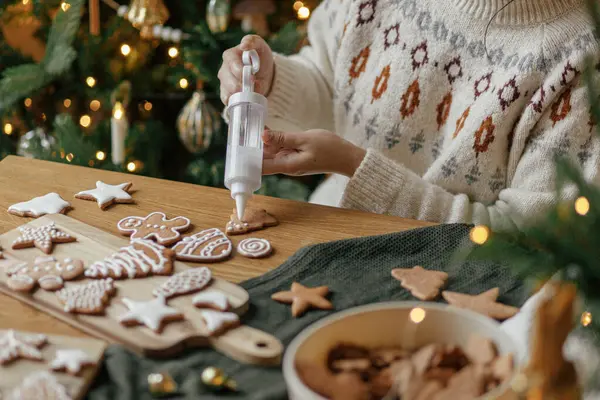 Hands decorating gingerbread cookies with icing on rustic wooden table on background of christmas golden lights. Atmospheric Christmas holiday traditions. Decorating cookies with sugar frosting