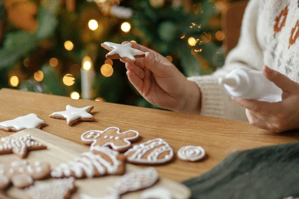 Hands decorating gingerbread cookies with icing on rustic wooden table on background of christmas golden lights. Atmospheric Christmas holiday traditions. Decorating cookies with sugar frosting