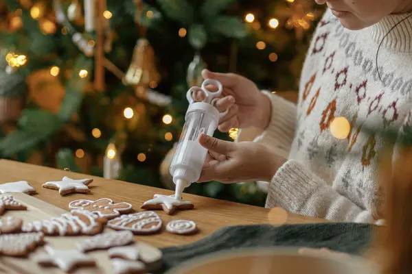 Decorating gingerbread cookies with icing on rustic wooden table at christmas tree golden lights. Atmospheric Christmas holiday traditions. Woman decorating cookies with sugar frosting