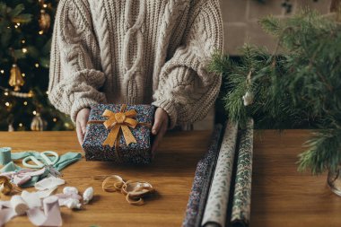 Samimi süveterli bir kadın elinde şık bir Noel hediyesi, kurdeleli, fiyonklu, ahşap masadaki klasik süslemeli bir paket kağıdıyla. Atmosferik kış tatili, Noel hediyelerini paketlemek.