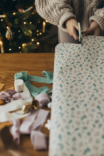 Wrapping christmas gifts. Hands in cozy sweater holding stylish festive wrapping paper with ribbons, vintage ornaments, bows on wooden table. Atmospheric winter holidays