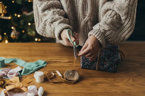 Wrapping christmas gifts. Hands in cozy sweater wrapping stylish present in festive wrapping paper with ribbons, vintage ornaments, bows on wooden table. Atmospheric winter holidays