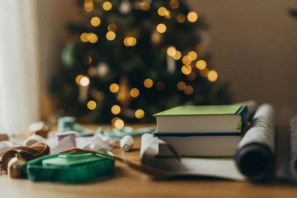 Wrapping christmas gifts. Stylish books as present in festive wrapping paper, ribbons, vintage ornaments on wooden table on background of christmas tree lights. Atmospheric winter holidays