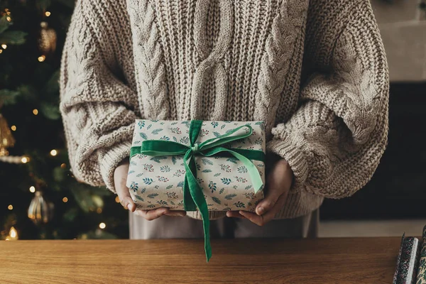 メリークリスマス スタイリッシュなクリスマスプレゼントを保持する居心地の良いセーターの女性 弓で紙を包むお祝いの贈り物 木のテーブルにリボン 大気の冬の休日 — ストック写真