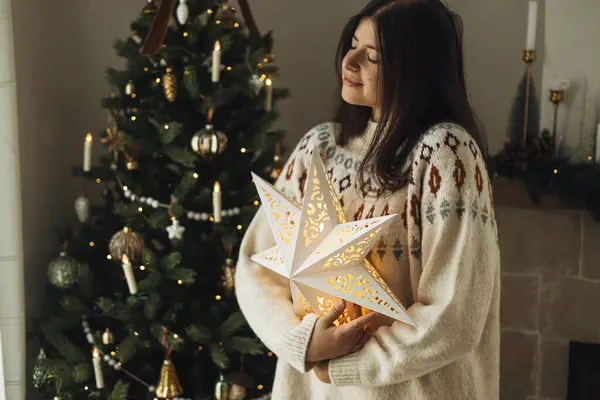 圣诞快乐 穿着舒适毛衣的女人拿着灯火通明的圣诞星 背靠装饰过的圣诞树 还有老式的浴盆和彩灯 大气中的圣诞节前夕 — 图库照片