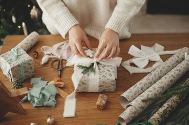 Bir kadın modern dekore edilmiş İskandinav odasında süslü süslemelerle süslü ahşap masaya kurdele ve köknar dalı ile şık bir Noel hediyesini paketliyor. Mutlu Noeller! Eller toplanıyor. Yakın çekim.