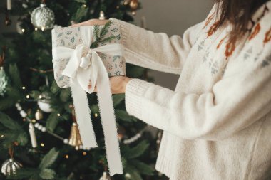 Mutlu Noeller ve mutlu Noeller! Elinde kurdele ve köknar dallarıyla şık bir Noel hediyesi tutan kadın, İskandinav odasındaki modern dekore edilmiş ağacın arka planına yakından bakıyor..