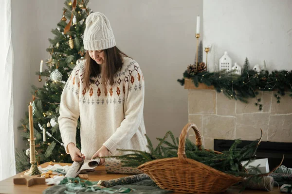 穿着舒适毛衣 头戴礼帽 头戴纸巾 头戴圣诞礼物的女人 坐在木制桌子上 装饰着节日装饰品 在装饰华丽的鸟房里 圣诞快乐 手拿着礼物 — 图库照片