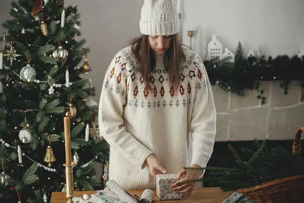 身穿时髦毛衣 头戴礼帽 头戴纸巾 头戴圣诞礼物的女人 坐在木制桌子上 房间里装饰着节日装饰品 圣诞快乐 手拿着礼物 — 图库照片