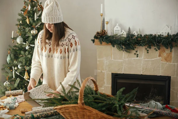 穿着舒适毛衣 头戴礼帽 头戴纸巾 头戴圣诞礼物的女人 坐在木制桌子上 装饰着节日装饰品 在装饰华丽的鸟房里 圣诞快乐 手拿着礼物 — 图库照片
