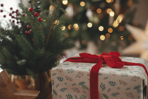 Счастливого Рождества и счастливых праздников! Стильный завернутый рождественский подарок и еловые ветки на столе против золотого света в праздничной скандинавской комнате. Атмосферное изображение