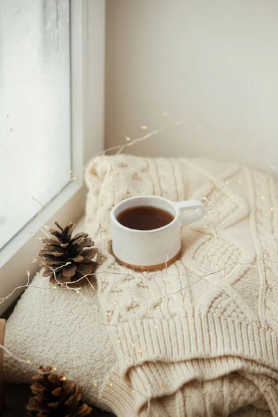 Cozy winter still life. Stylish cup of tea with cozy knitted sweater, pine cone, wooden star and golden lights on windowsill in festive scandinavian room. Autumn and christmas holidays