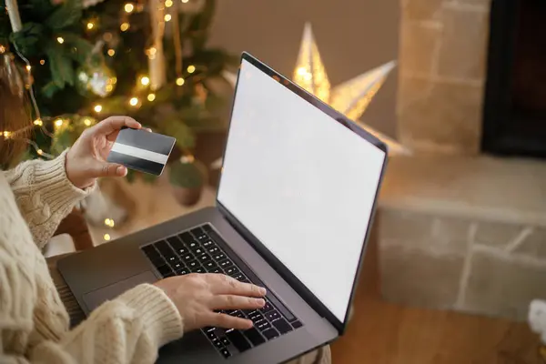 Christmas shopping online and black friday sales. Woman hands holding credit card on background of laptop with blank screen in festive decorated christmas room with lights. Space for text