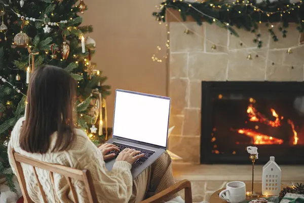 Woman in cozy sweater working on laptop with empty screen on background of christmas tree lights in stylish festive decorated room. Christmas shopping online and remote work template