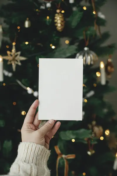 クリスマスカードモックアップ 金色のライトが付いているスタイリッシュな装飾されたクリスマス ツリーの背景に空の挨拶カードを握る手 テキストのためのスペース 季節の挨拶のテンプレートとヴィンテージの装飾 — ストック写真