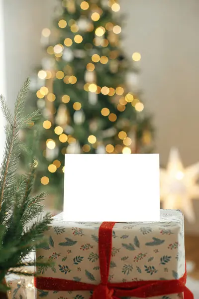 Christmas card mock up. Empty greeting card stylish wrapped christmas gift against festive christmas tree with golden lights. Space for text. Season greetings postcard template