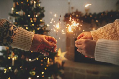 Mutlu yıllar! Modern şömine karşısında yanan havai fişekleri tutan eller ve altın ışıklı Noel ağacı. Arkadaşlar ve aile ellerinde yanan maytaplarla, atmosferik arifeyle kutlama yapıyorlar.