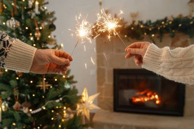 Mutlu yıllar! Modern şömine karşısında yanan havai fişekleri tutan eller ve altın ışıklı Noel ağacı. Arkadaşlar ve aile ellerinde yanan maytaplarla, atmosferik arifeyle kutlama yapıyorlar.