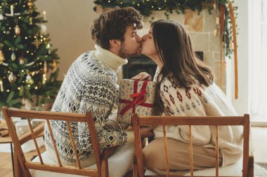 Mutlu, genç bir aile rahat kazaklar içinde şöminenin arka planında şık Noel hediyelerini değiş tokuş ederken modern manto ve ışıklandırmalı noel ağacı. İyi tatiller.!
