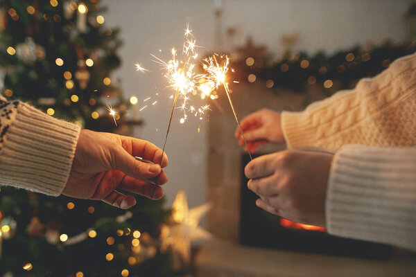 Hands holding burning fireworks against modern fireplace and christmas tree with golden lights. Happy New Year! Friends and family celebrating with burning sparklers in hands, atmospheric eve