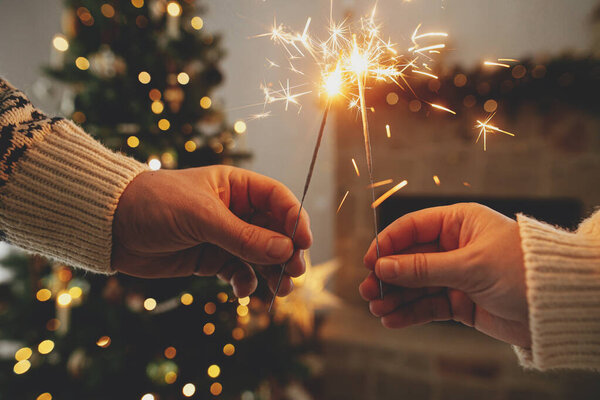 С Новым Годом! Горящие искры в руках на фоне современного загородного камина и рождественской елки с золотыми огнями. Фейерверк светится в руках, пара празднует в праздничном украшенном помещении