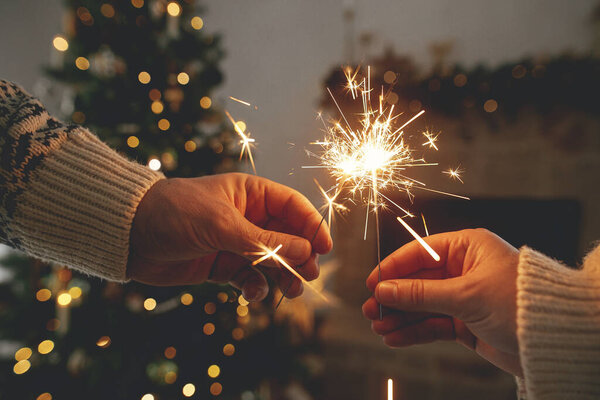 С Новым Годом! Горящие искры в руках на фоне современного загородного камина и рождественской елки с золотыми огнями. Фейерверк светится в руках, пара празднует в праздничном украшенном помещении