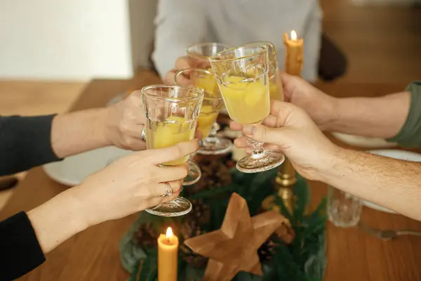 Hands with mimosa cocktail glasses clicking on background of stylish christmas table setting with fir branches, pine cones and candles. Friends and family celebrating and toasting, happy holidays